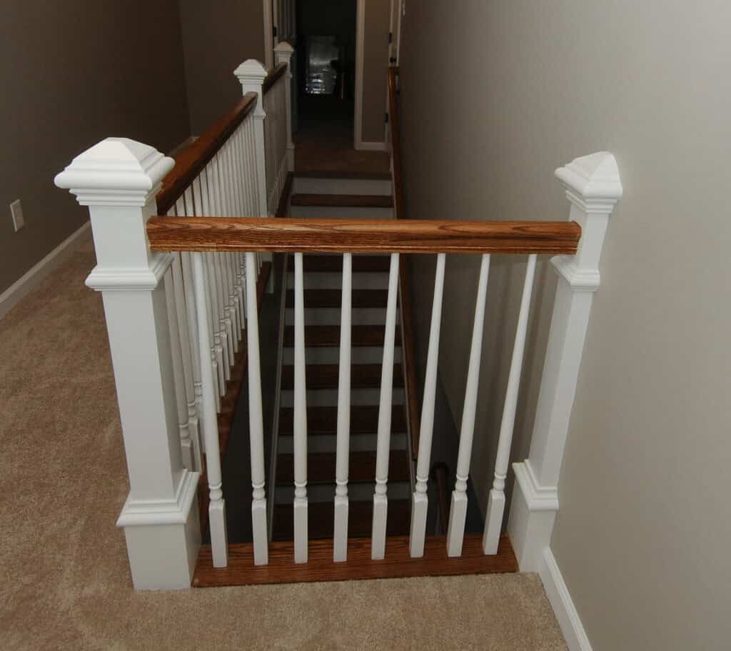 Wooden stair railing with white spindles and a dark wood handrail at the top of a carpeted staircase in a hallway with beige walls. The stairs lead down to a lower floor.