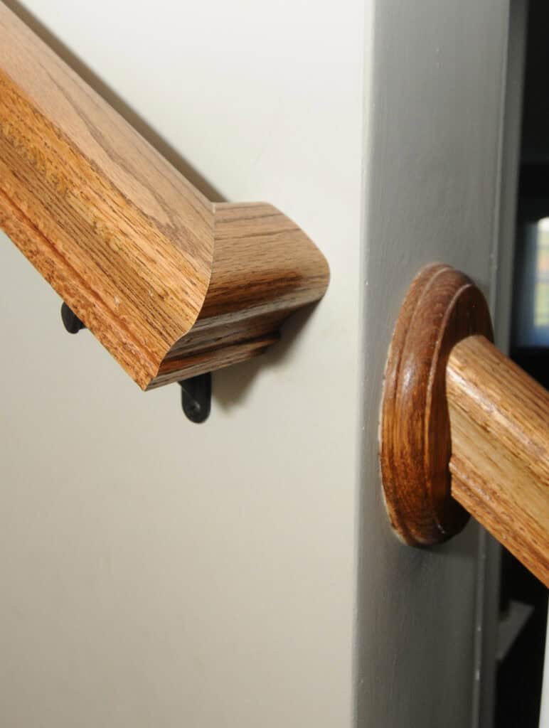 Close-up of a staircase handrail made of finished wood, mounted on a light-colored wall. The handrail ends with a decorative angled cut, and is supported by black metal brackets.