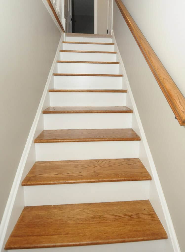 A straight staircase with light wooden steps and white risers, flanked by beige walls and a wooden handrail on the right side, leading up to a hallway.