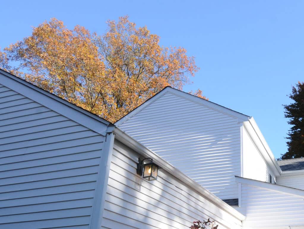 White-sided houses with angled roofs are shown under a clear blue sky. Sunlight casts sharp shadows on the walls, and autumn trees with orange and yellow leaves rise in the background.