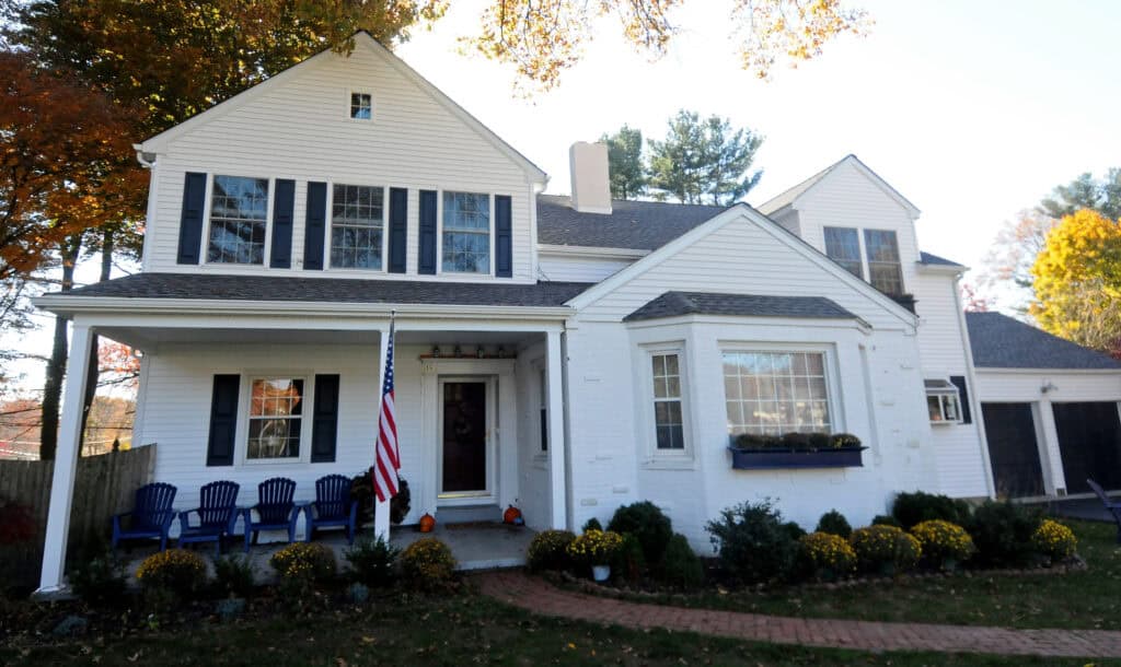 A white two-story house with black shutters, a front porch, several blue chairs, an American flag, and a garden with yellow flowers and shrubs. Trees with autumn leaves are visible in the background.