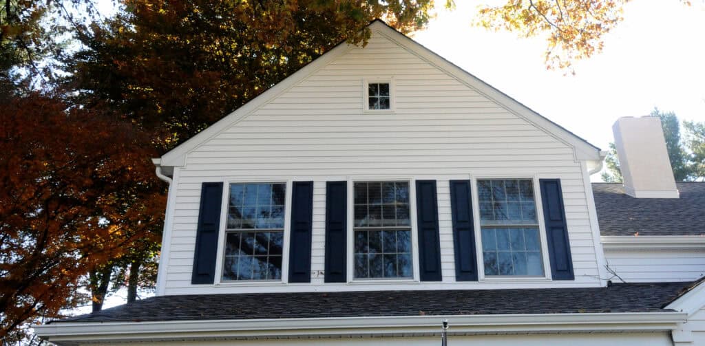 The upper portion of a white house with three large windows, each flanked by black shutters. There is a small square window above, and trees with autumn leaves are visible in the background.