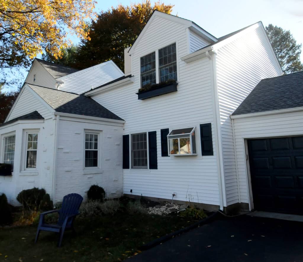 A white two-story house with black shutters, an attached garage, and a driveway. There is a blue chair on the lawn and trees with autumn-colored leaves in the background.