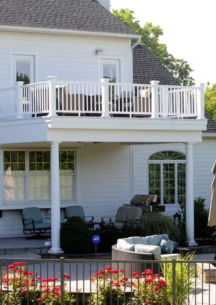 A white house with a second-floor balcony featuring a white railing, a covered patio area below with outdoor furniture, a grill, green shrubs, and red flowers in the foreground.