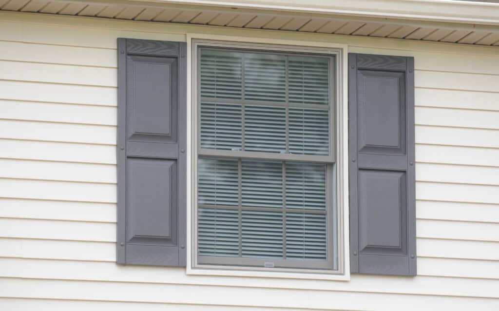 A window with closed gray shutters on both sides, set in a cream-colored vinyl siding wall. The window has white trim and horizontal blinds are visible through the glass.