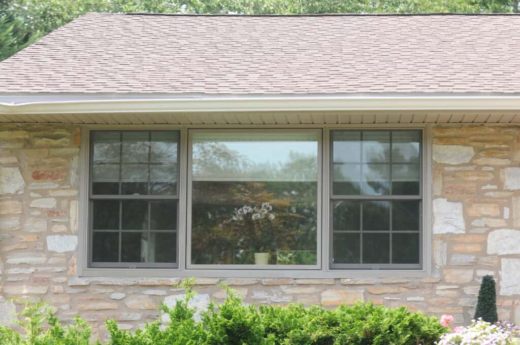 A large rectangular window with three glass panes set in a stone wall of a house, with shrubs in front. Reflections of trees are visible in the window, and roof shingles are above.