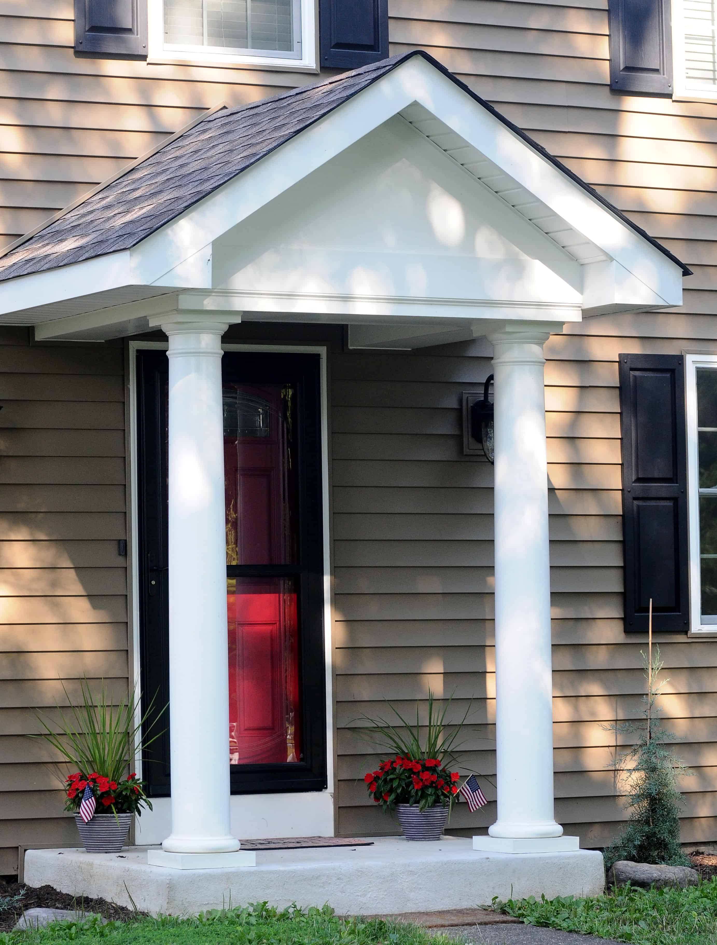 A house entrance with tan siding, white trim, black shutters, a red door, white columns supporting a small portico, and two planters with red flowers and mini American flags on either side of the door.