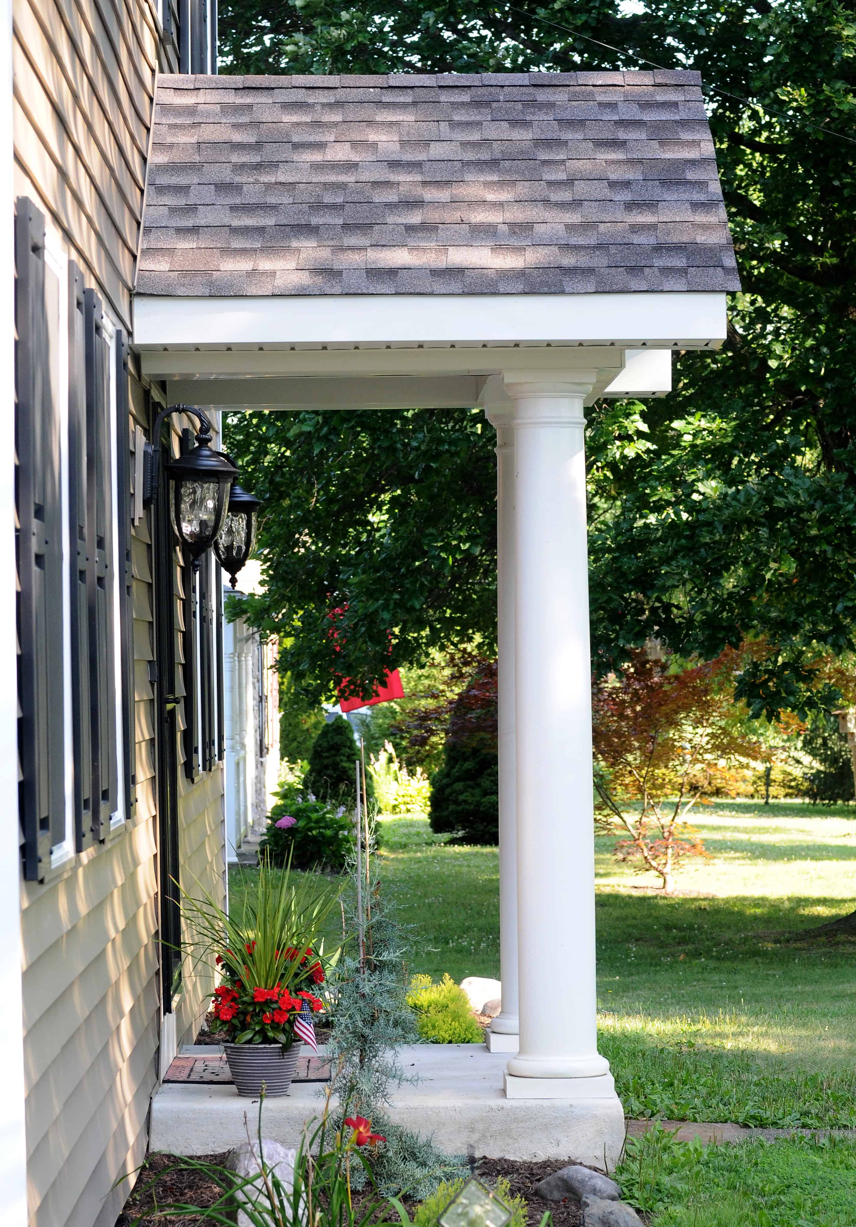 A white column supports a small porch roof by the side of a house with tan siding. Flower pots and plants line the porch, and a green lawn and trees are visible in the background.