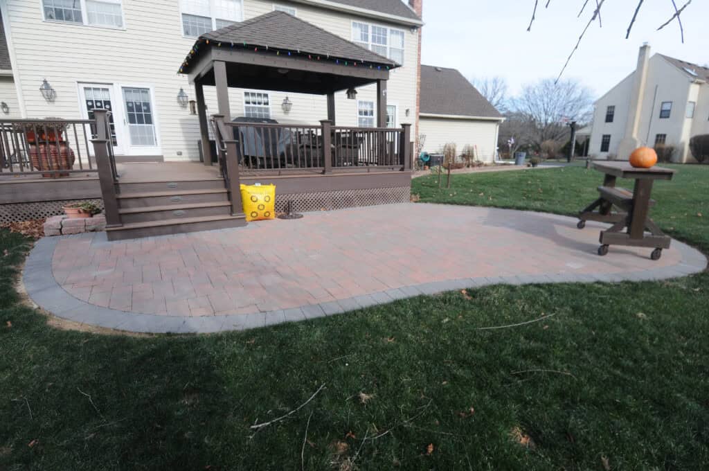 A backyard patio with red and gray pavers, a wooden deck with stairs, and a roofed section decorated with string lights. A yellow block sits by the stairs and a wooden table with a pumpkin is on the patio. Grass surrounds the area.