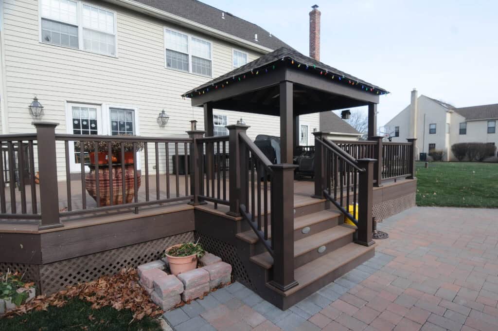 A spacious backyard deck attached to a house, featuring brown railings, a covered gazebo with string lights, stairs leading down to a paved patio, and potted plants near the steps.