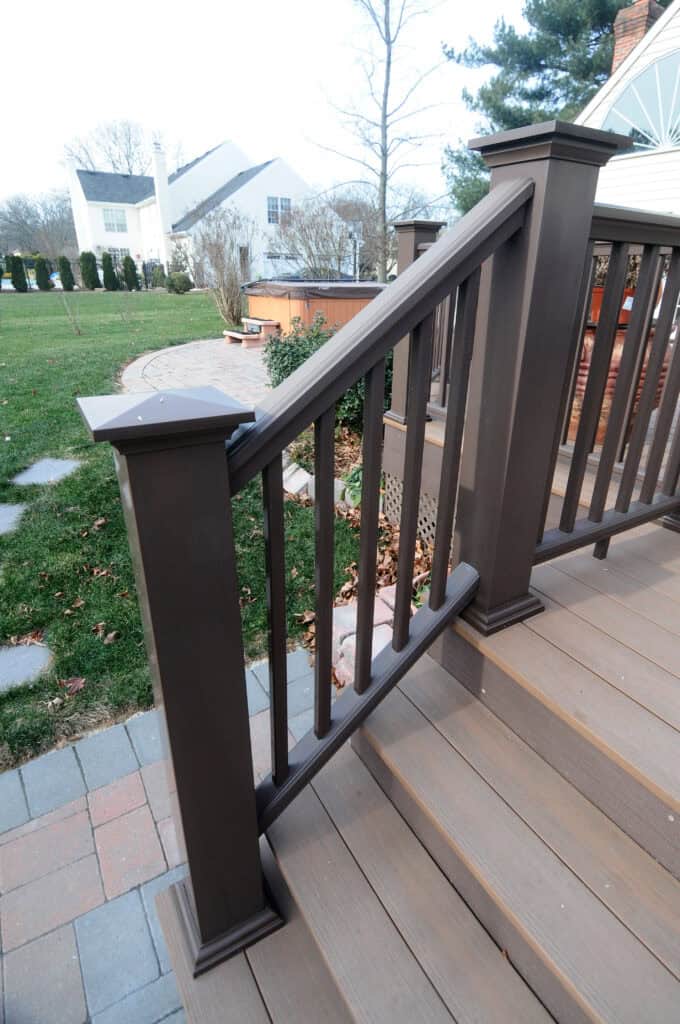 A brown wooden deck stair railing with vertical balusters, leading down to a patio with stone pavers. Green grass and shrubs are visible in the background along with white houses and trees.