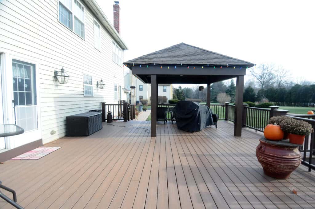 A spacious backyard deck with brown flooring, a covered pergola, a grill covered with a black tarp, outdoor furniture, and large planters with pumpkins. The deck is attached to a white house and overlooks a green lawn.