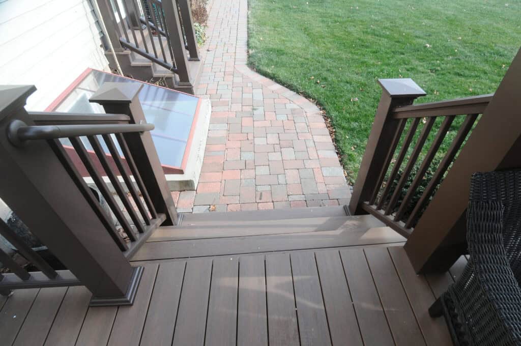 View from the top of brown wooden stairs with black railings, leading down to a red and gray brick walkway, with green grass and a window well visible to the left.