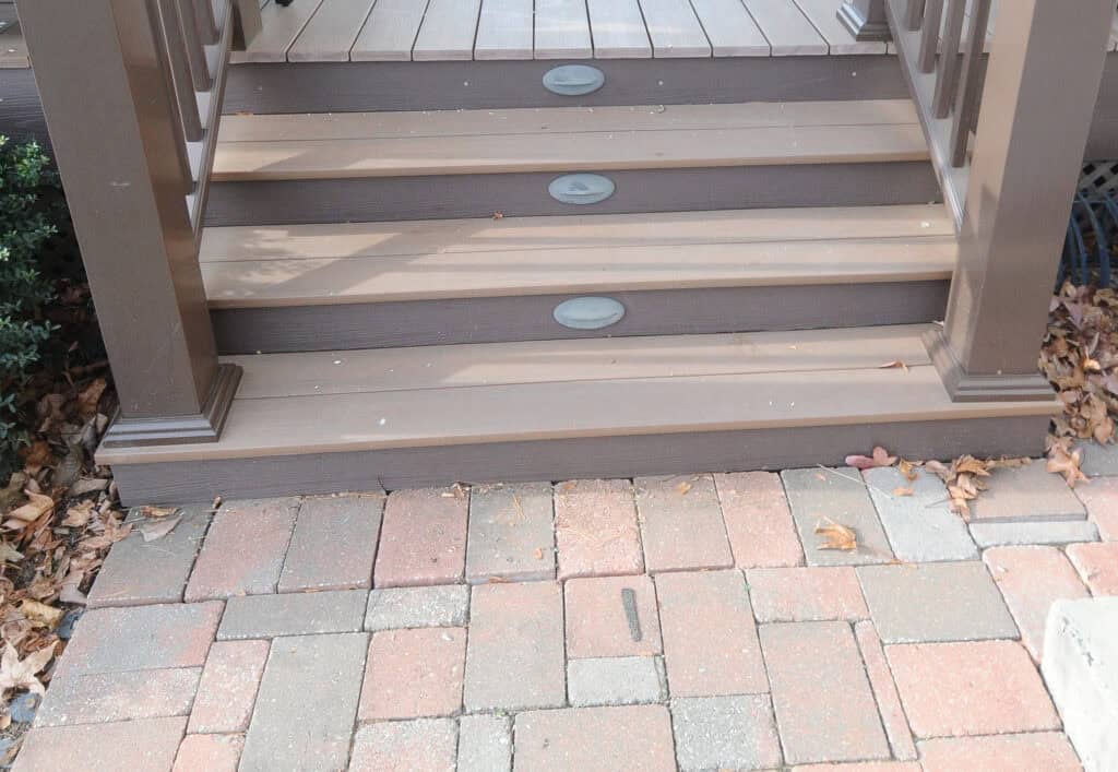 Three brown wooden steps with oval lights are attached to a deck, leading down to a patio made of red, brown, and tan bricks. Fallen leaves are scattered along the edges of the steps and patio.