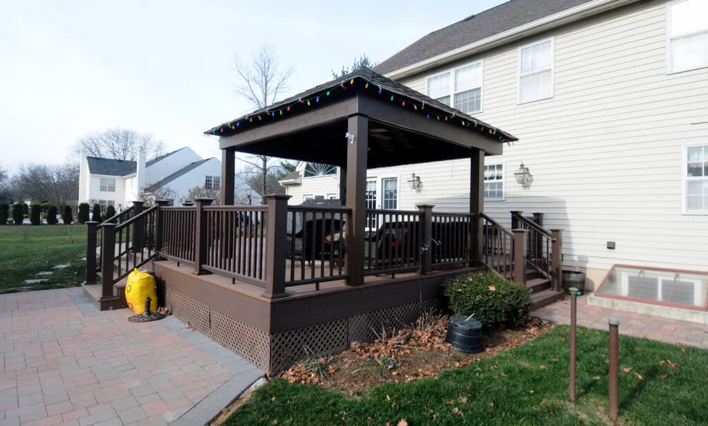 A wooden deck with a gazebo and string lights is attached to the back of a light-colored house, surrounded by a yard, some shrubs, and a paved patio area.
