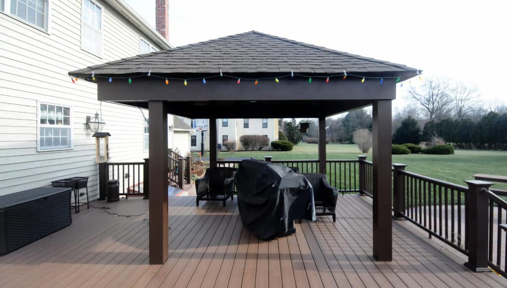 A backyard wooden gazebo with a shingled roof decorated with multicolored string lights, covering a grill and patio furniture, on a spacious deck attached to a house. A large lawn and trees are visible in the background.