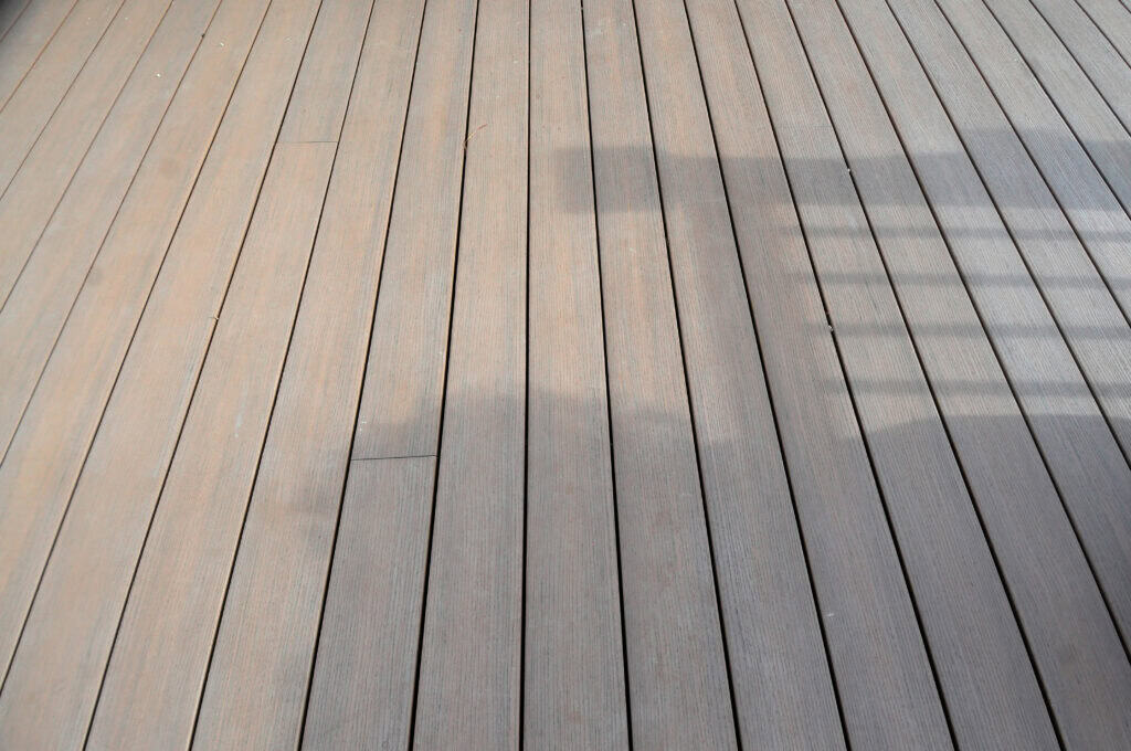 Light brown wooden deck boards with a shadow of a railing and balusters cast across part of the surface, creating a geometric pattern. The decking appears clean and evenly spaced.