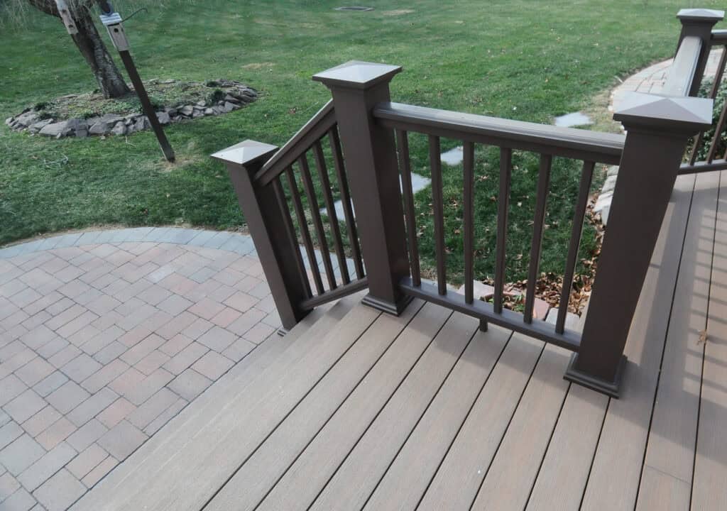 A close-up view of a wooden deck with brown railings and stairs leading down to a patio with light brown paving stones; green grass and a tree are visible in the background.