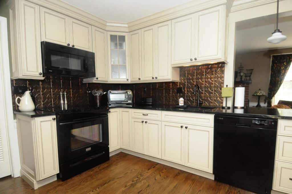 A modern kitchen with cream-colored cabinets, black appliances, a dark metallic backsplash, wooden floor, and various countertop items including a toaster oven, kettle, utensils, and paper towels.