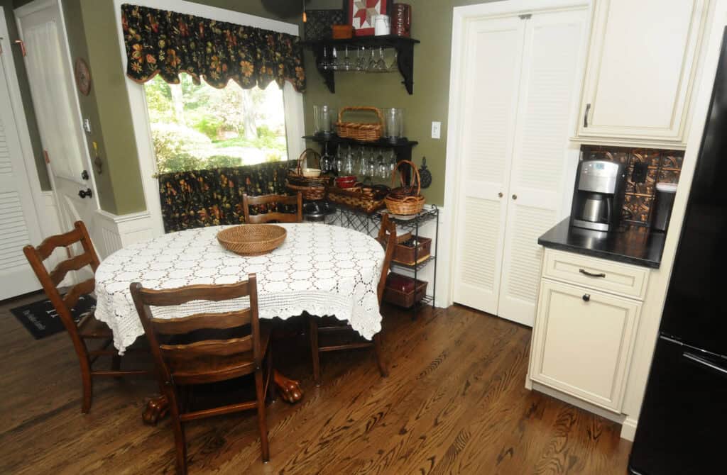 A cozy kitchen with a round table covered by a lace tablecloth, four wooden chairs, baskets on shelves, white cabinets, and a coffee maker on a black countertop. A window with floral curtains lets in natural light.