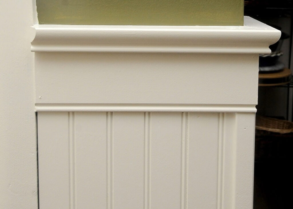 Close-up of a white, decorative wainscoting panel with vertical grooves, topped with an ornate molding and ledge, against a light-colored wall.