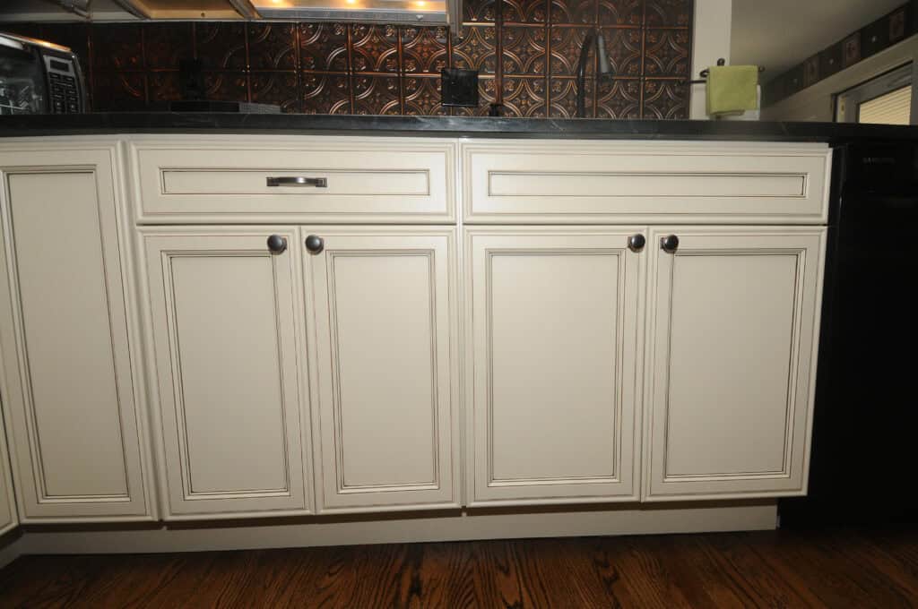 White kitchen cabinets with decorative trim and black handles, set against a dark countertop and patterned metallic backsplash on a wood floor.