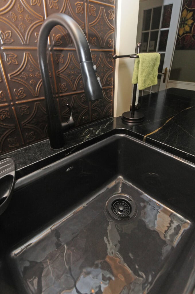 A black kitchen sink with a modern black faucet, set against a decorative brown tiled backsplash. A green towel hangs on a stand next to the sink on a dark countertop.