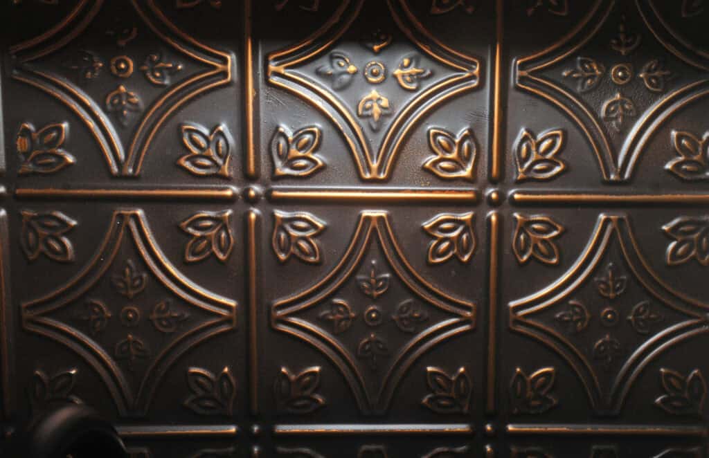 Embossed metal tile with a repeating geometric and floral pattern, featuring raised floral and diamond shapes. The metal has a dark bronze or copper finish with subtle highlights.