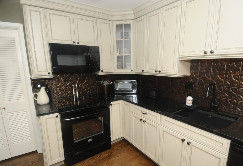 A modern kitchen with cream-colored cabinets, black countertops, a black stove, microwave, sink, toaster oven, kettle, and patterned dark backsplash on wooden floors.