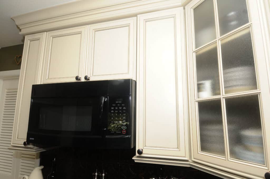 A black microwave oven is built into cream-colored kitchen cabinets, with a section of frosted glass cabinet doors revealing stacked dishes inside.
