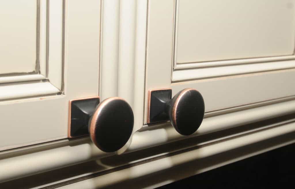 Close-up of two cream-colored cabinet doors with round, dark metal knobs featuring a subtle bronze edge, set against a grooved and paneled surface.