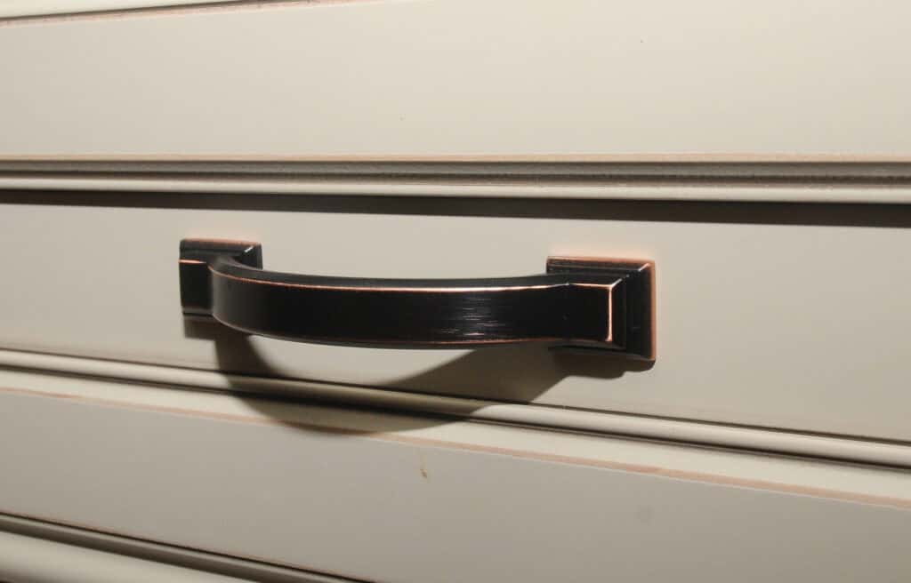 A close-up of a dark metal drawer handle attached to a cream-colored drawer with decorative grooves.