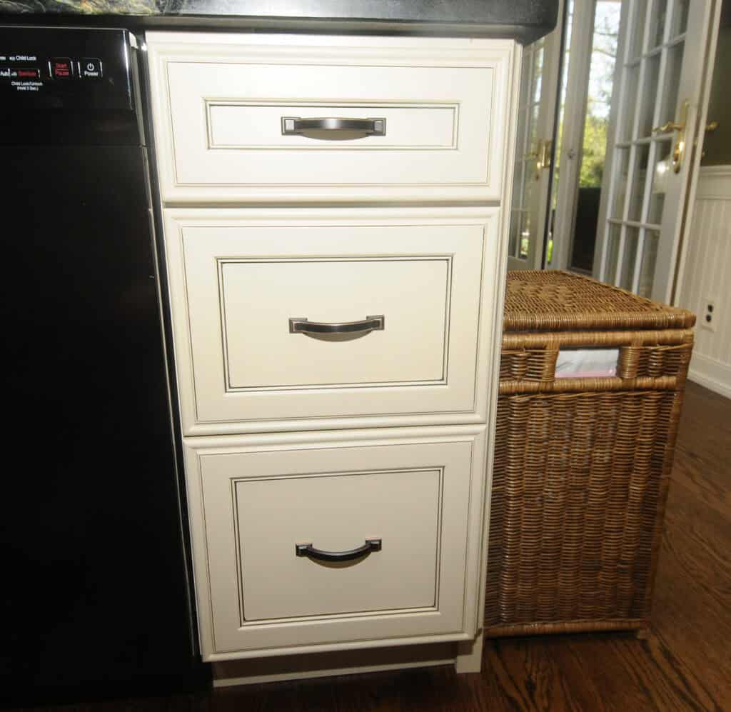 Three cream-colored kitchen drawers with dark handles are set beside a black dishwasher and a woven wicker basket hamper on a wooden floor, with windows and a door in the background.