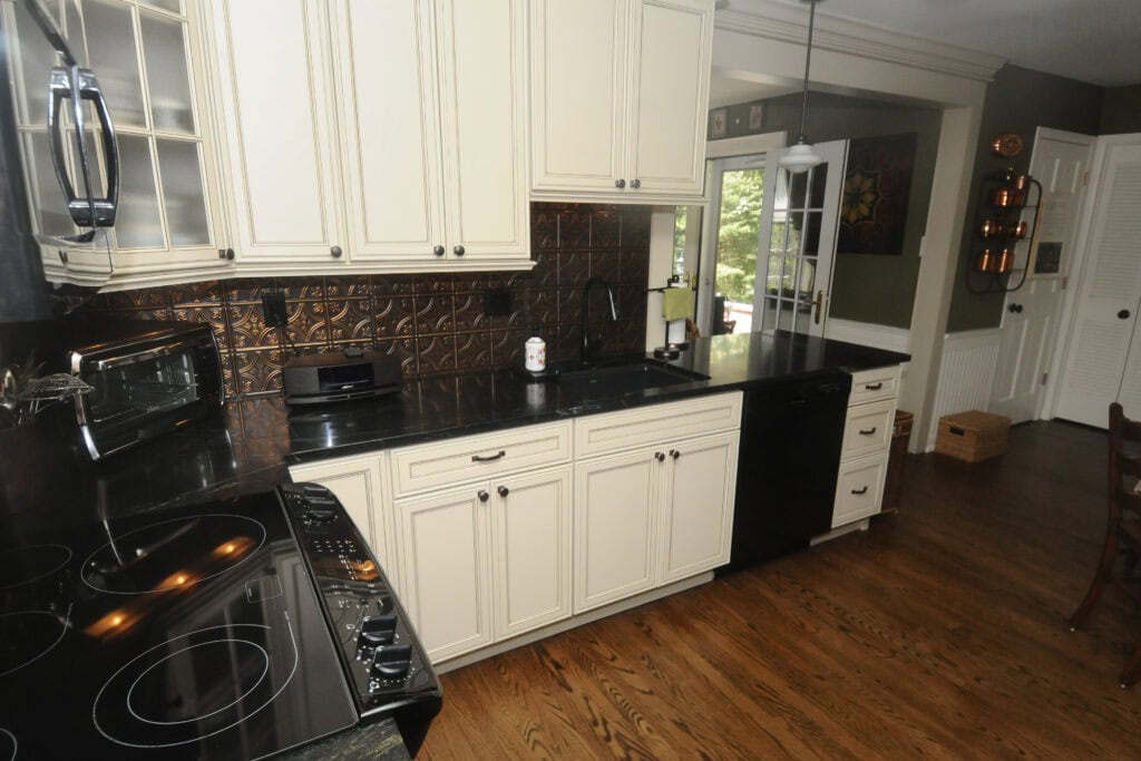A kitchen with cream-colored cabinets, black countertops, a black electric stove, microwave, dishwasher, toaster oven, and patterned metallic backsplash, with wooden flooring and sunlight coming through a window.