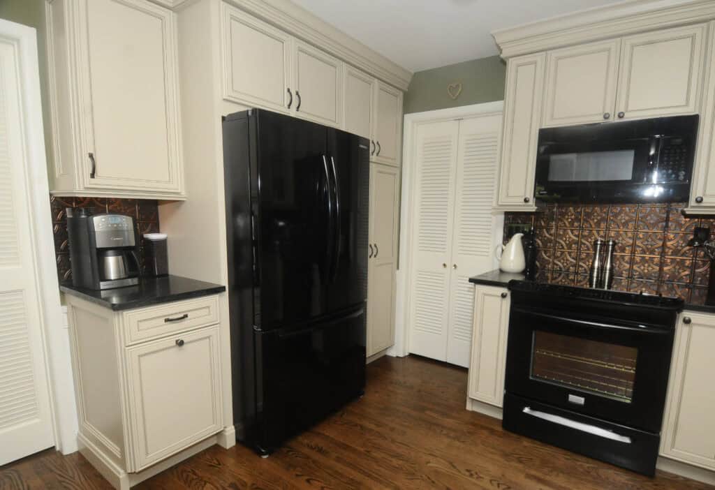 Modern kitchen with cream-colored cabinets, black appliances including a refrigerator, oven, and microwave, a coffee maker on the counter, and wood flooring. Copper backsplash adds a warm accent behind the counters.