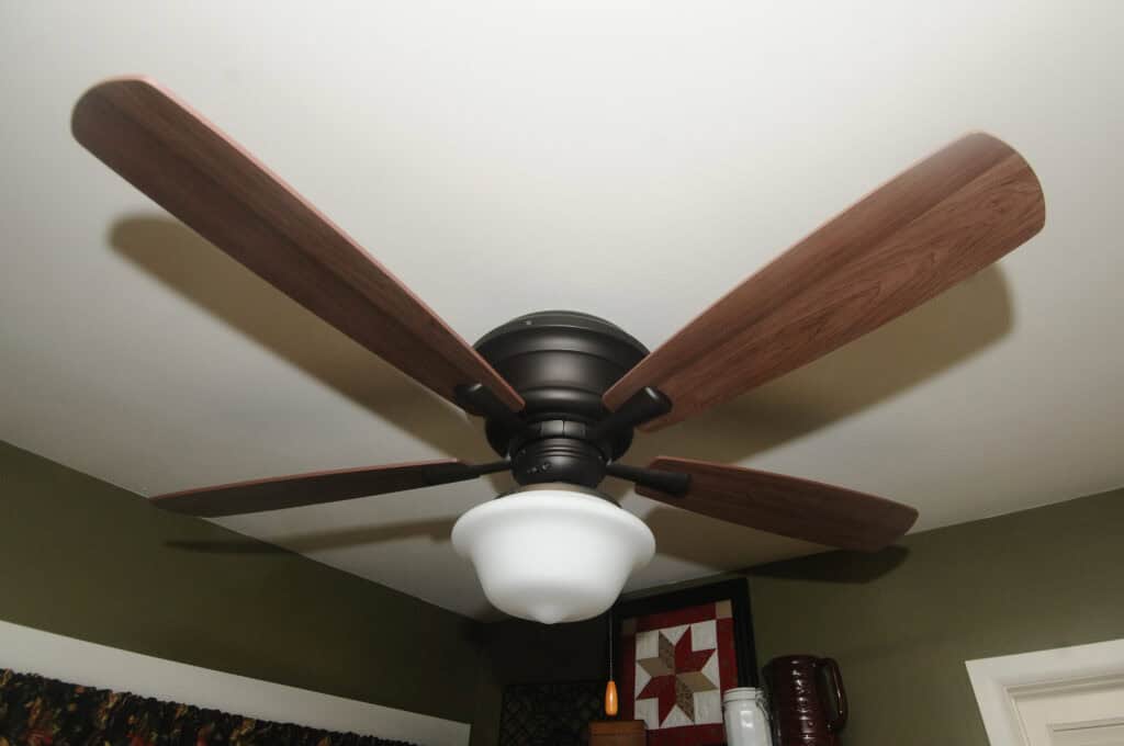 A ceiling fan with five wooden blades and a frosted glass light fixture is mounted on a white ceiling in a room with green walls and framed decor.
