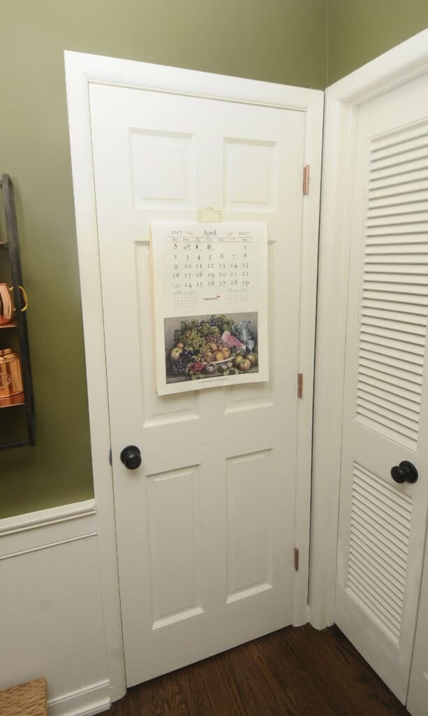 A white door with a wall calendar hanging on it, displaying the month of April and a picture of assorted fruits and vegetables. The room has green walls, a white louvered door, and dark wood flooring.