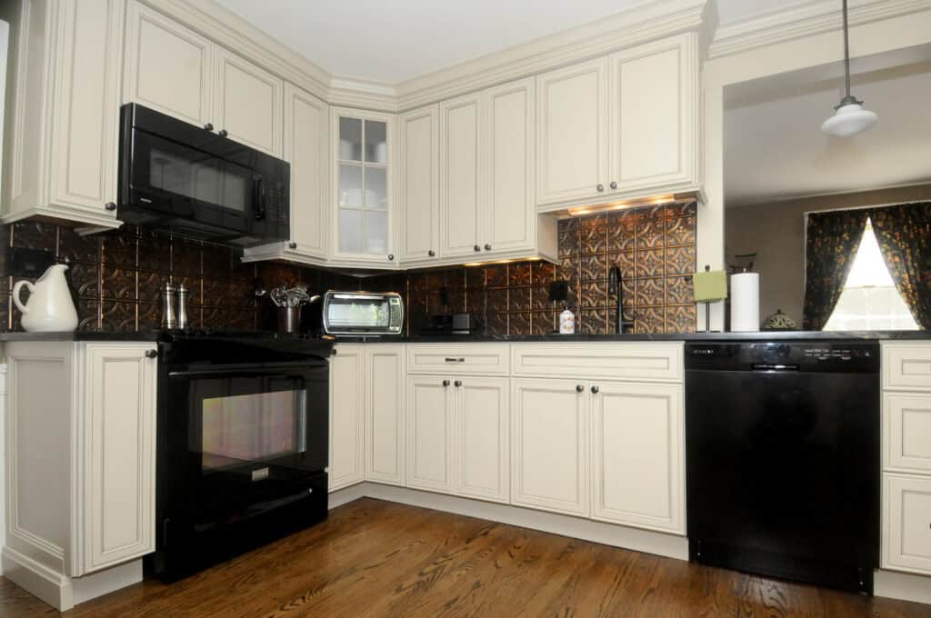 A modern kitchen with cream-colored cabinets, black appliances, a brown patterned backsplash, wood flooring, and a window with floral curtains in the adjoining room.