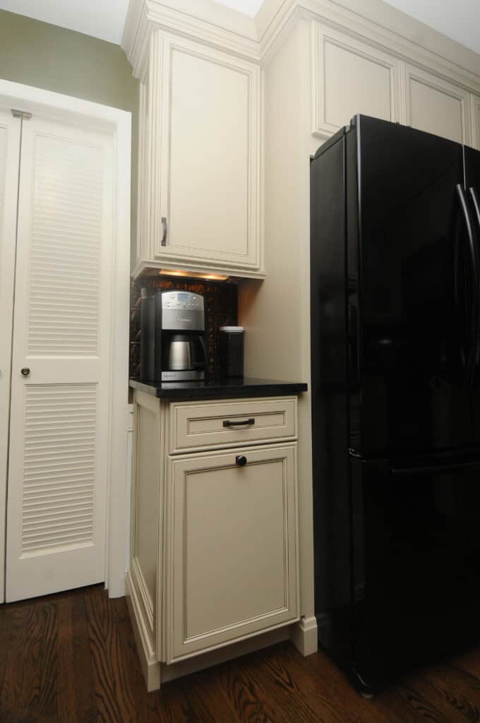 A modern kitchen corner with cream-colored cabinets, a black countertop, a coffee maker, a small metallic box, and a black refrigerator on dark wood flooring.