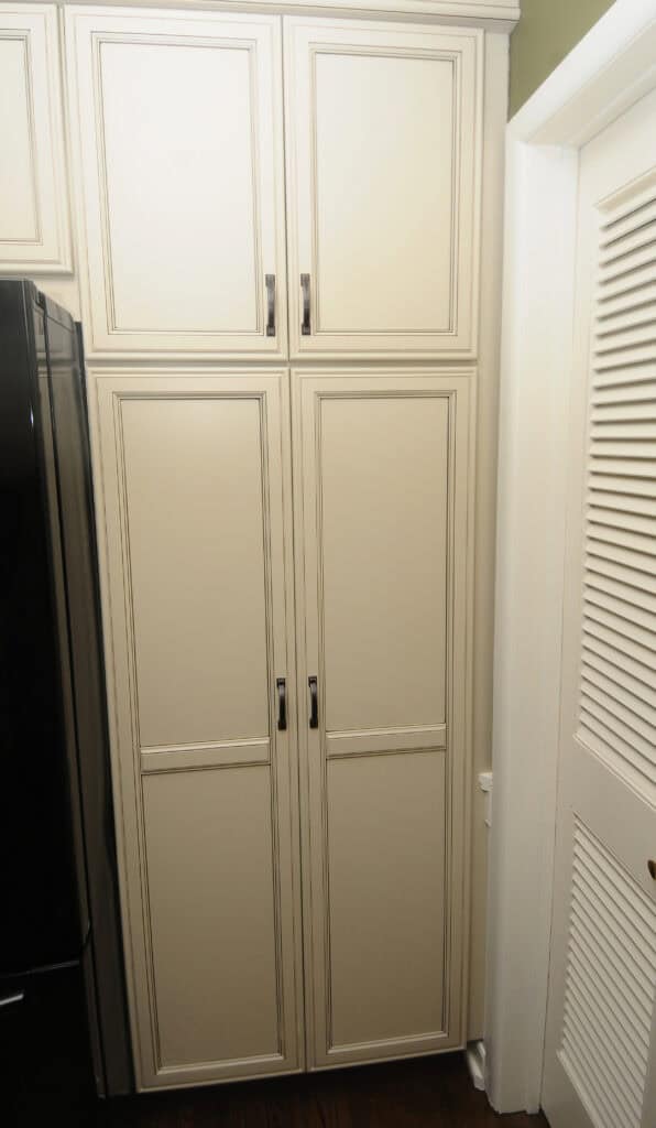 Tall, cream-colored kitchen pantry cabinet with four paneled doors and silver handles, positioned next to a louvered door and a black refrigerator.