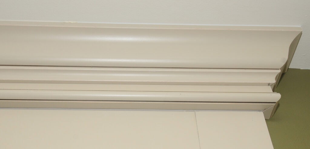 Close-up of beige crown molding installed at the corner where the wall meets the ceiling, with a portion of green-painted wall visible in the bottom right corner.