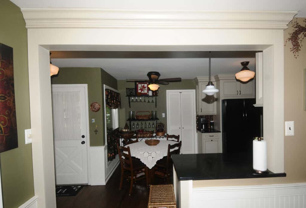 A cozy kitchen and dining area with a table covered by a lace tablecloth, wooden chairs, white cabinets, black appliances, and warm lighting. The walls are green, and there are decorative items on shelves and the door.