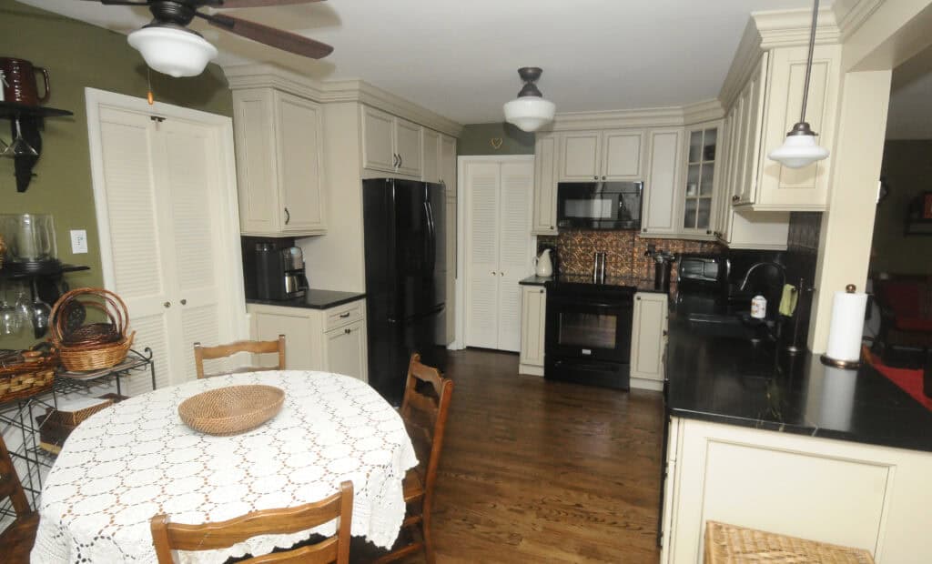 A kitchen with cream cabinets, black appliances, and dark countertops. A round table with a lace tablecloth and a wicker basket is in the foreground, surrounded by wooden chairs. The floor is wooden, and the walls are green.