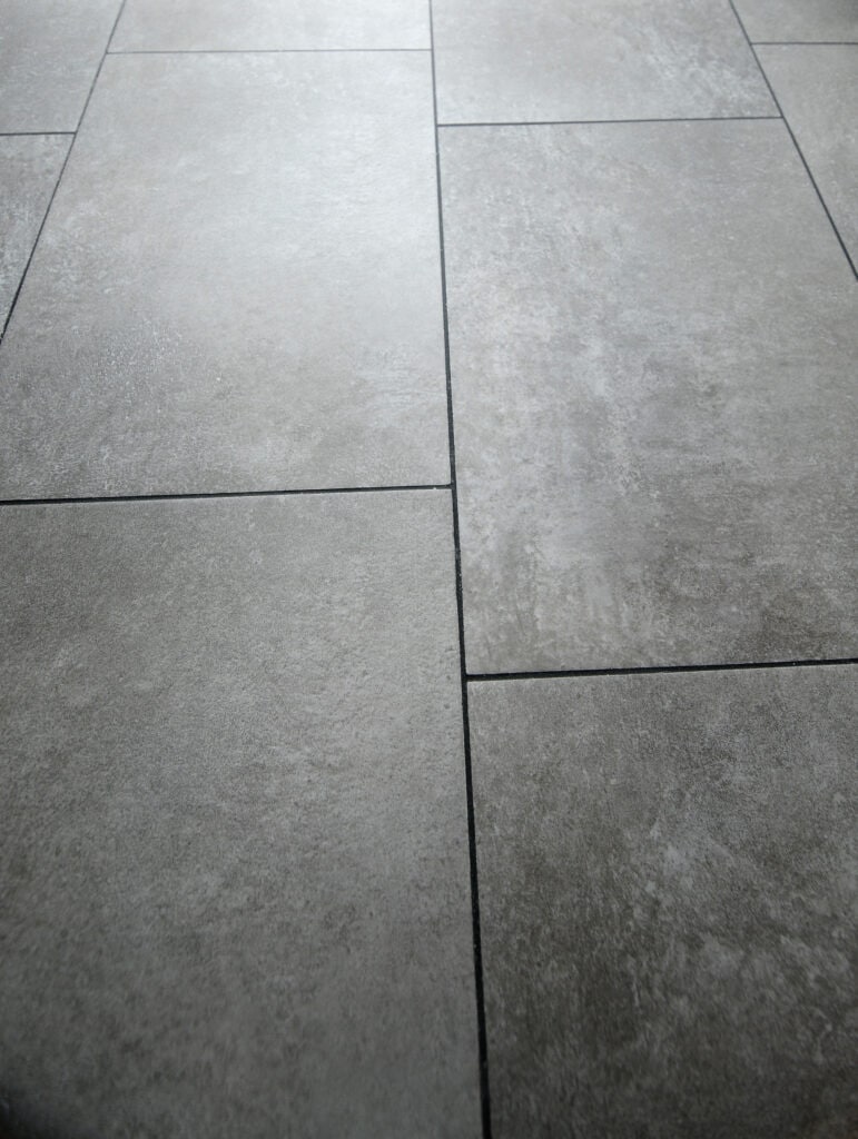 Close-up view of gray ceramic floor tiles with a matte finish, arranged in a staggered pattern. The grout lines between the tiles are visible.
