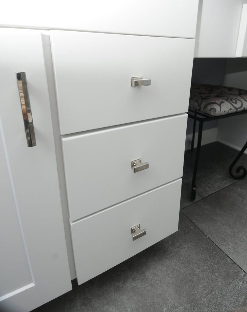 Three white drawers with modern silver handles are shown next to a cabinet door, also with a silver handle. The floor is dark gray tile, and part of a cushioned bench with a patterned seat is visible on the right.