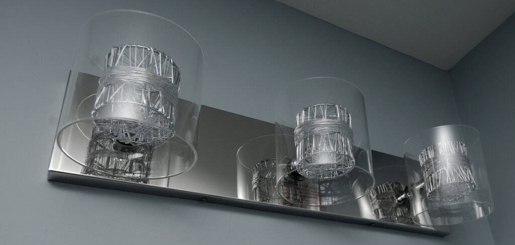 A modern wall-mounted light fixture with four cylindrical glass covers and metallic wire accents, attached to a rectangular metal base, against a light blue wall.