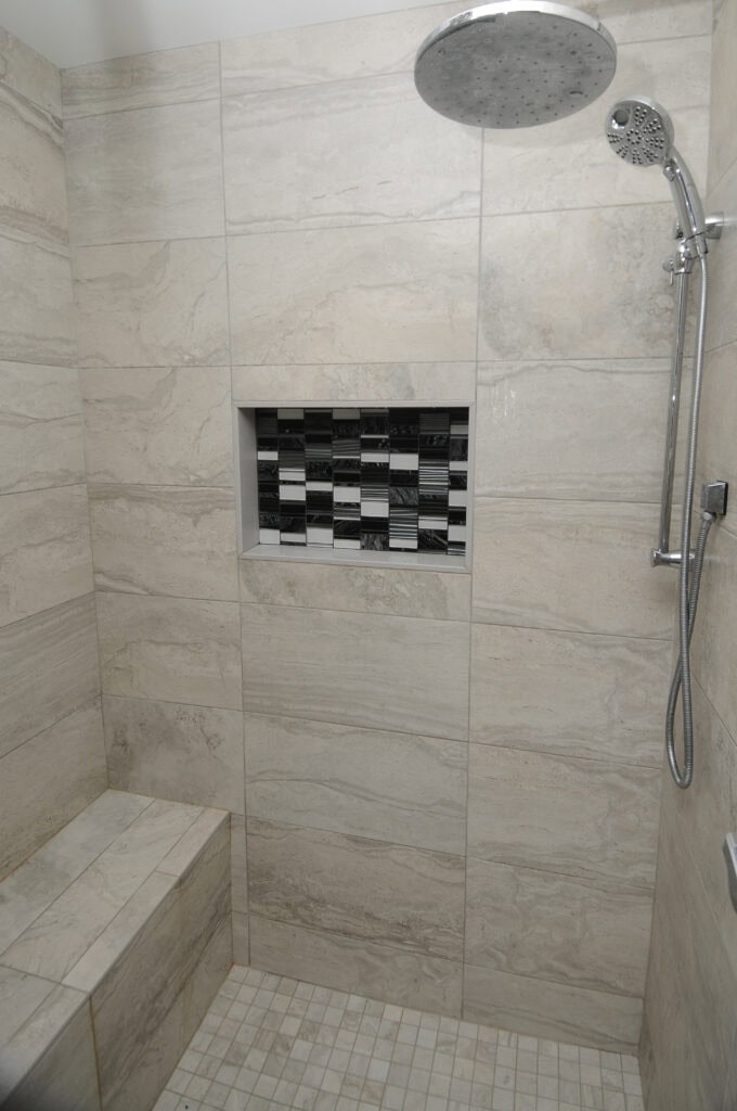 Modern walk-in shower with large beige tiles, a built-in bench, a recessed shelf with black and white mosaic tiles, a rainfall showerhead, and a handheld shower attachment.