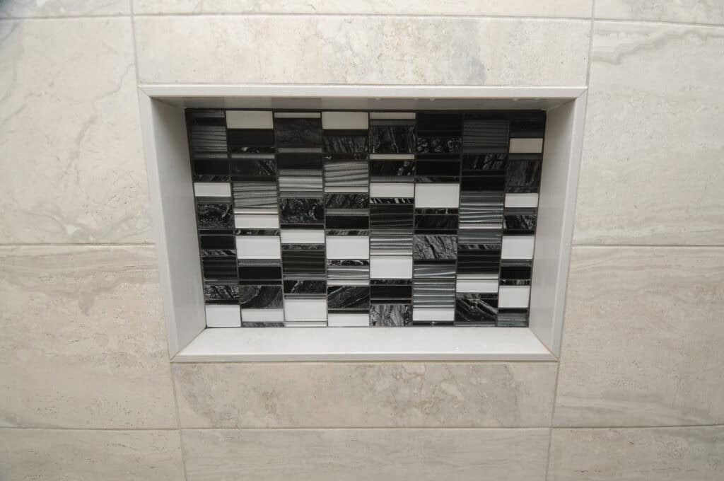 A recessed shower niche with a black, white, and gray rectangular mosaic tile pattern surrounded by large light gray tiles.