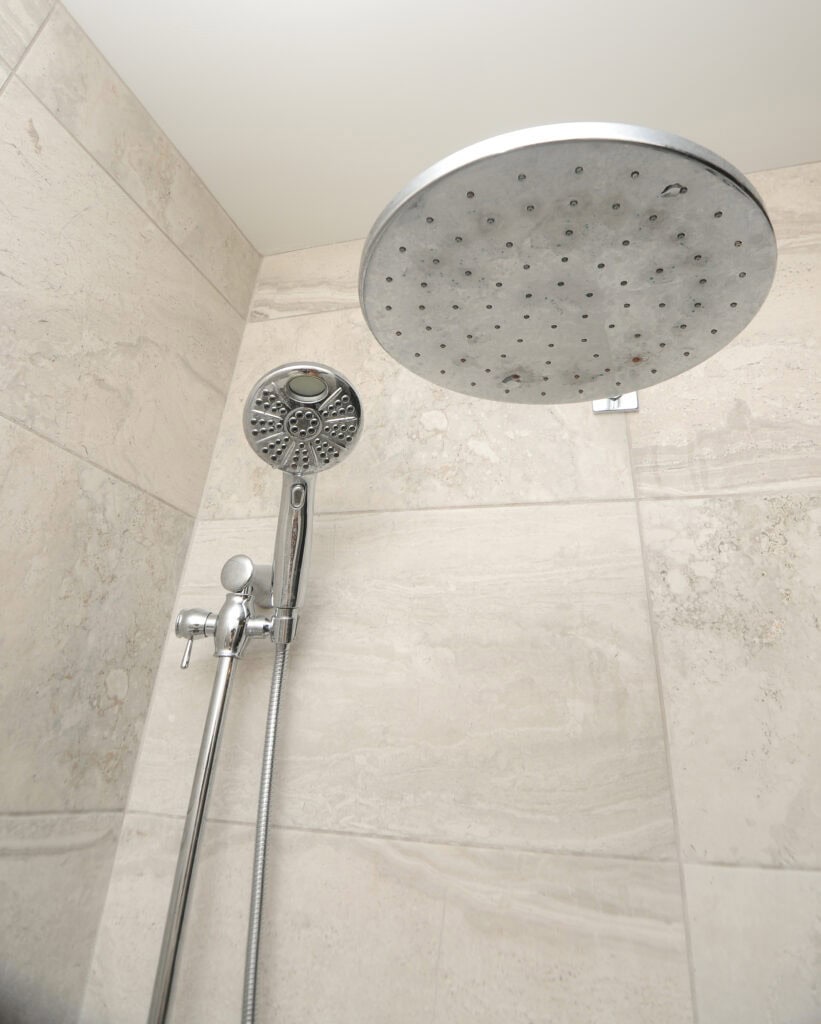 Modern shower with beige tiled walls, featuring a large round overhead rainfall showerhead and a handheld showerhead attached to a vertical bar with a flexible hose.