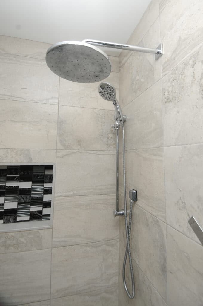Modern shower with large, round overhead rain showerhead and a smaller handheld showerhead, both attached to a tiled wall. A built-in shelf with black and white tiles is visible on the left.